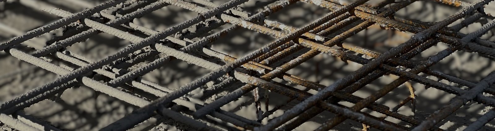 Das Bild zeigt ein engmaschiges Gitter aus rostigen, teilweise mit Zementresten bedeckten Stahlstäben. Die Stäbe sind rechtwinklig miteinander verschweißt und bilden eine typische Bewehrungsmatte, wie sie im Betonbau zur Verstärkung verwendet wird. Im Hintergrund und auf den Stäben sind Schattenmuster zu erkennen, die durch das Sonnenlicht entstehen.

                        